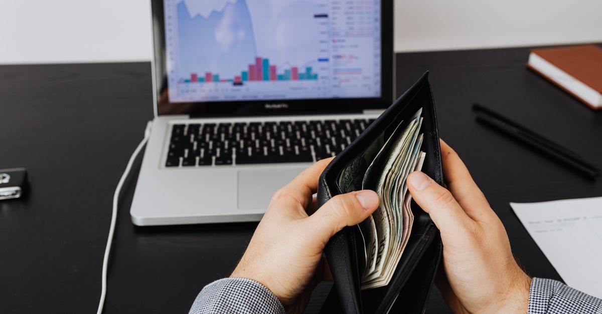 Man holding a leather wallet with bills in front of a laptop displaying financial graphs.
