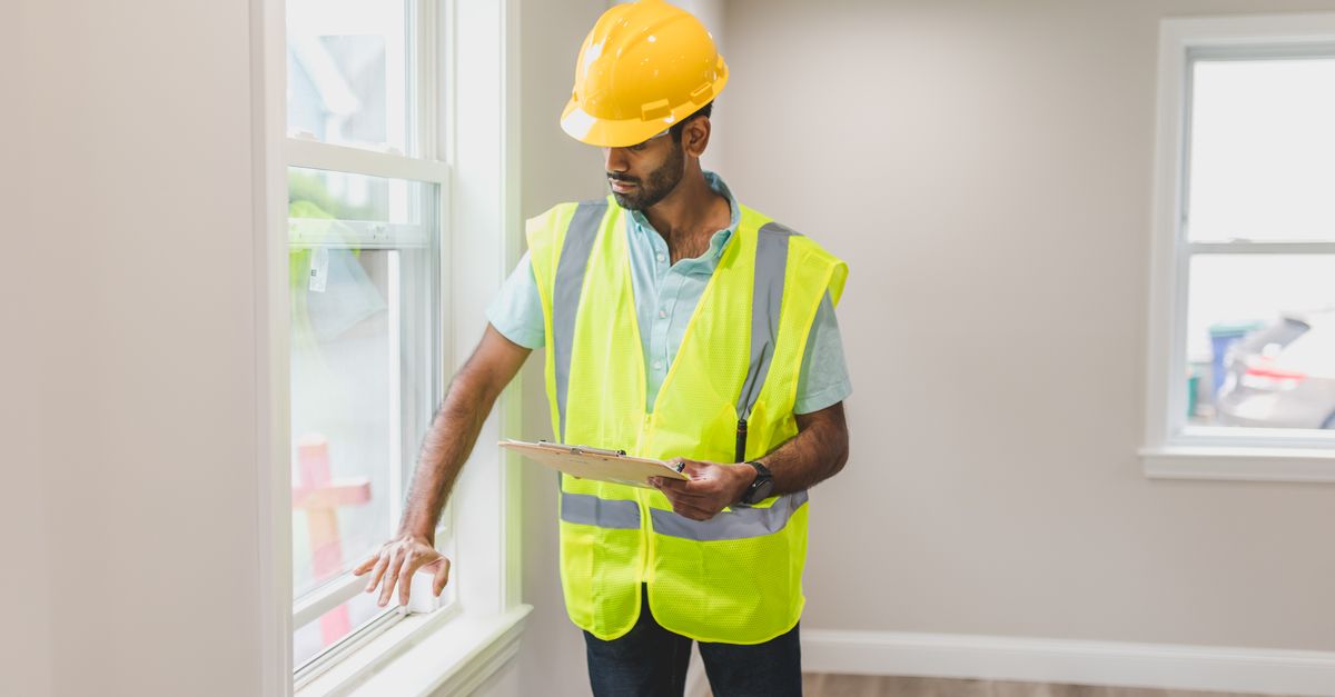 Real estate agent inspecting window indoors, wearing safety vest and hard hat, ensuring home safety.