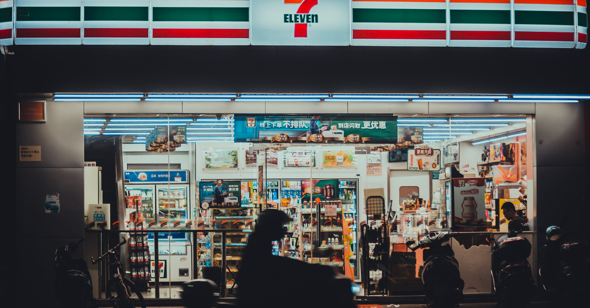 A vibrant 7-Eleven store front lit up during nighttime with a silhouette of a motorbike rider passing by.
