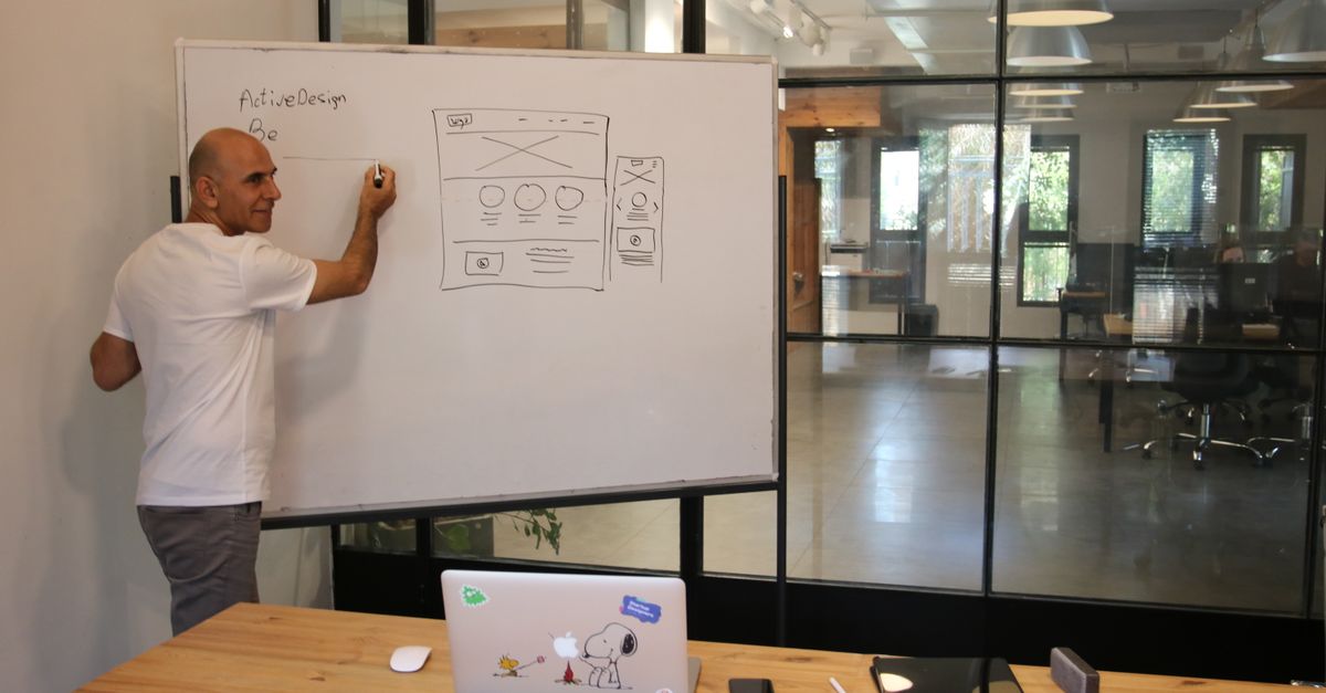 Side view of middle aged male leader in white t shirt writing on whiteboard during presentation in modern office space with glass walls