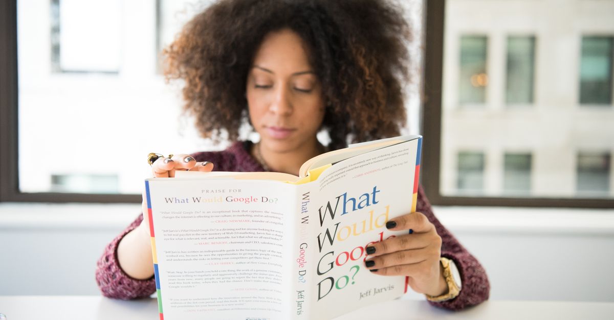 A woman reading 'What Would Google Do?' at a desk by a window in a modern office.
