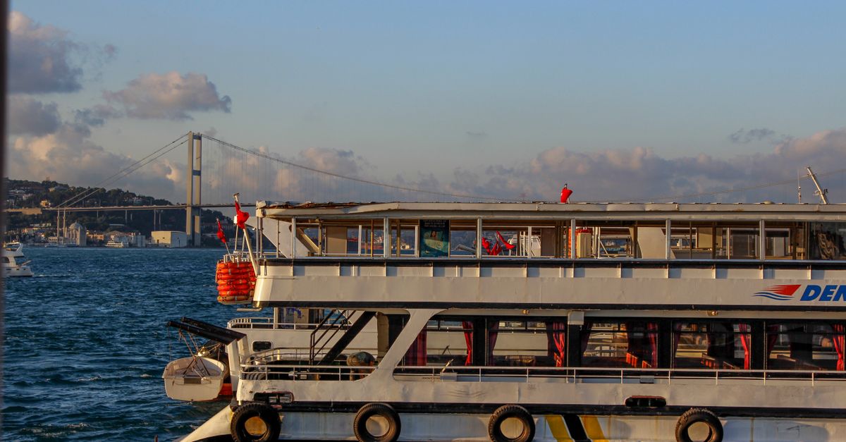 Scenic view of a ferry sailing on the Bosphorus in Istanbul, with the Fatih Sultan Mehmet Bridge in the background at sunset.