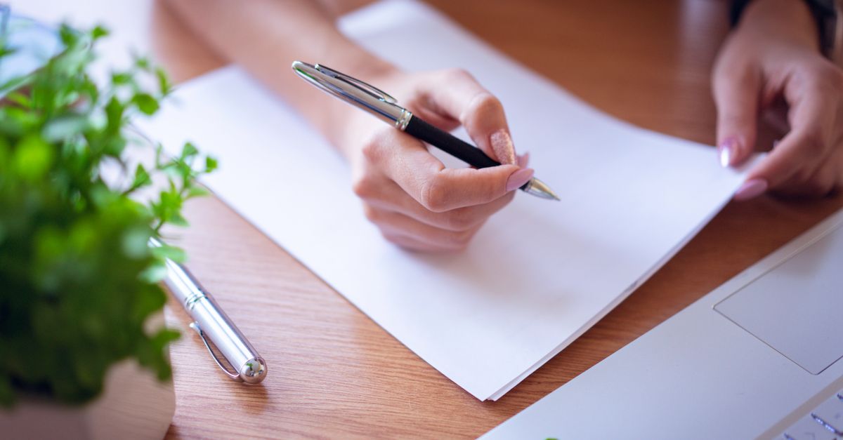 Close-up of a woman writing with a black pen on white paper next to a laptop and plant.