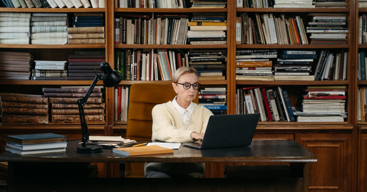Woman in a yellow sweater working on a laptop amidst books in a library setting.