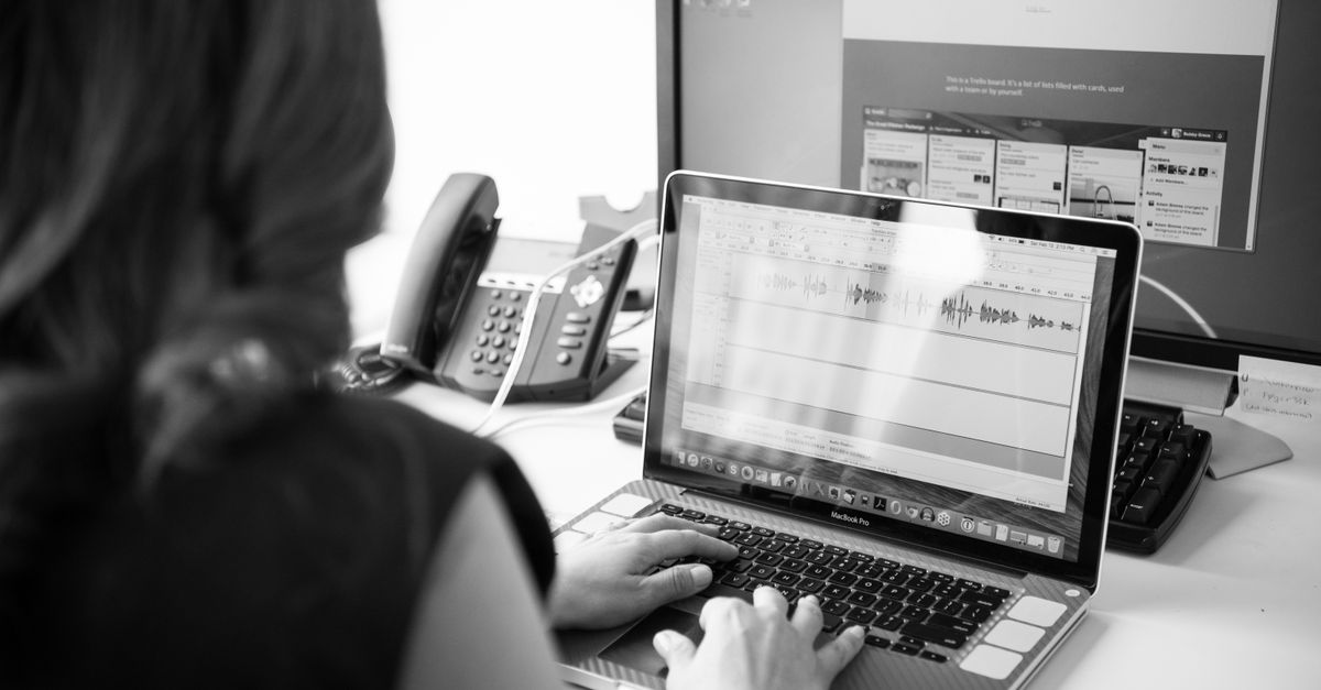 Black and white office scene with a woman working on a laptop in front of dual monitors.