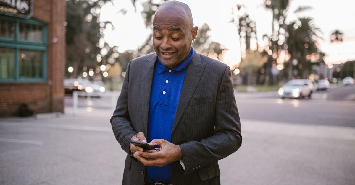 An adult man in a suit is happily using his smartphone outside during sunset, showcasing communication and modern lifestyle.