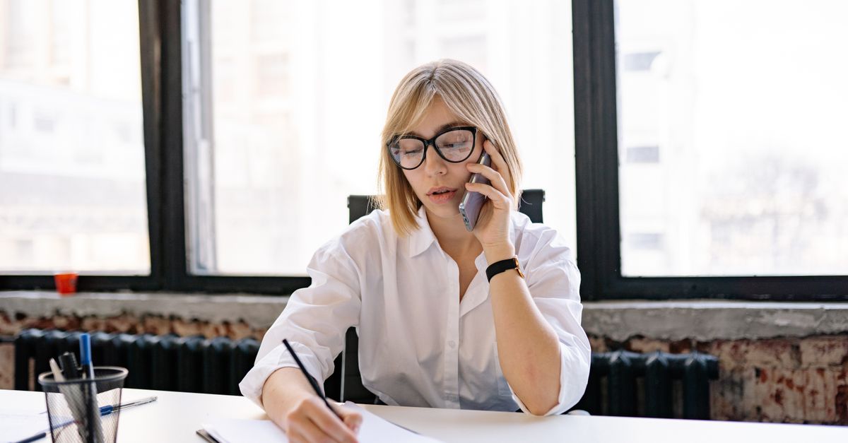 A woman in an office setting multitasking with a phone call and taking notes.