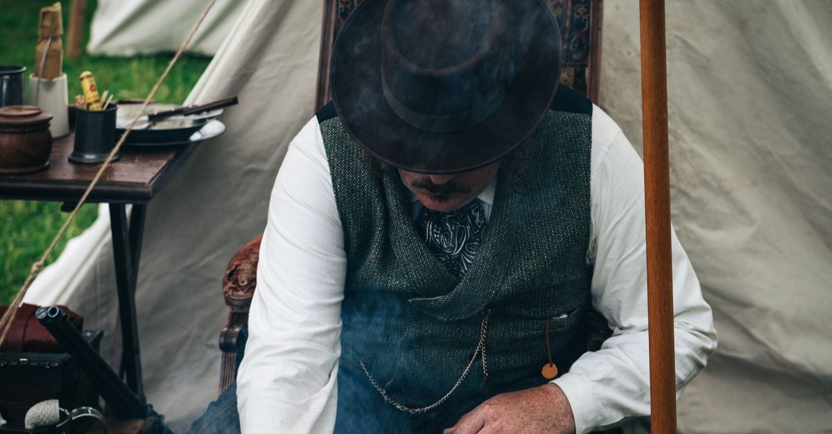 A man in vintage attire relaxes by a campsite with a smoking pipe.