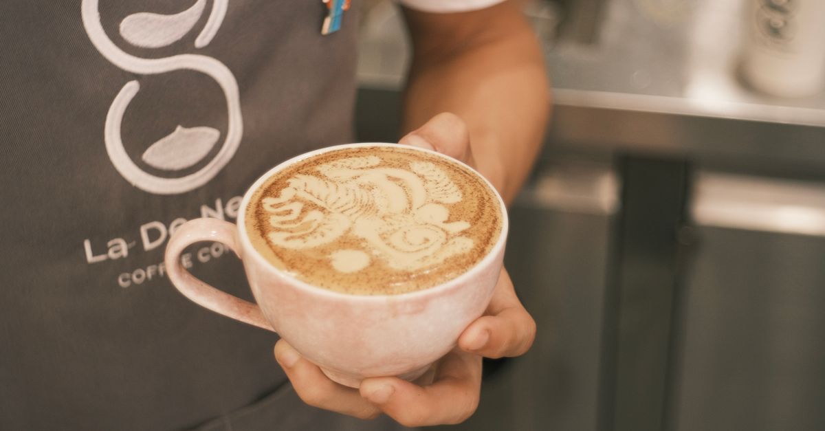 Close-up of a barista holding a cappuccino with intricate latte art, indoors in a coffee shop.