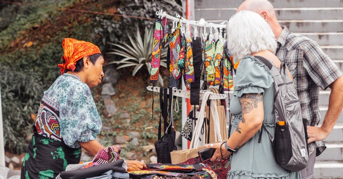 A local artisan selling colorful textiles to customers at a market stall outdoors.