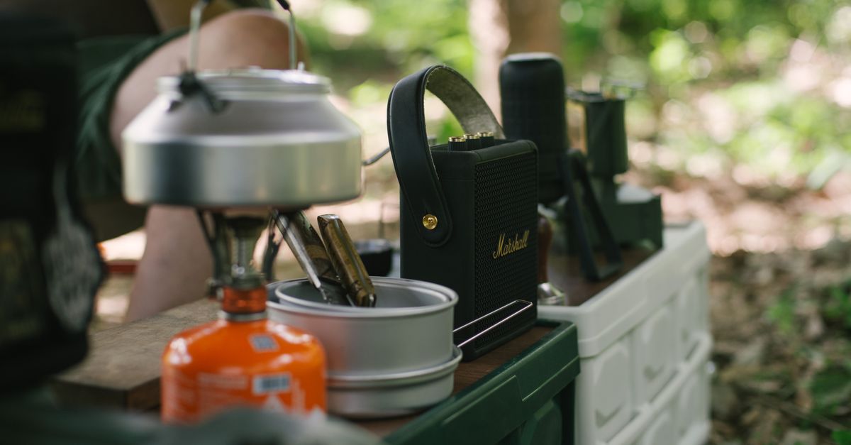 Outdoor camping gear including a gas stove and kettle on a table in a forest setting.