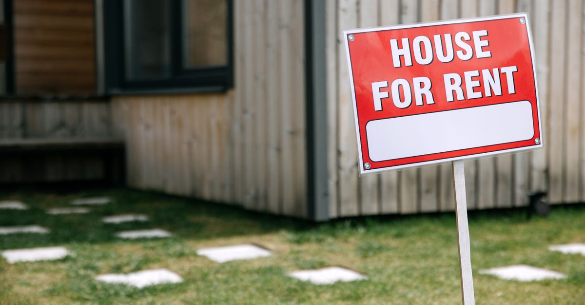 A modern wooden house with a prominent 'House for Rent' sign in the green yard.