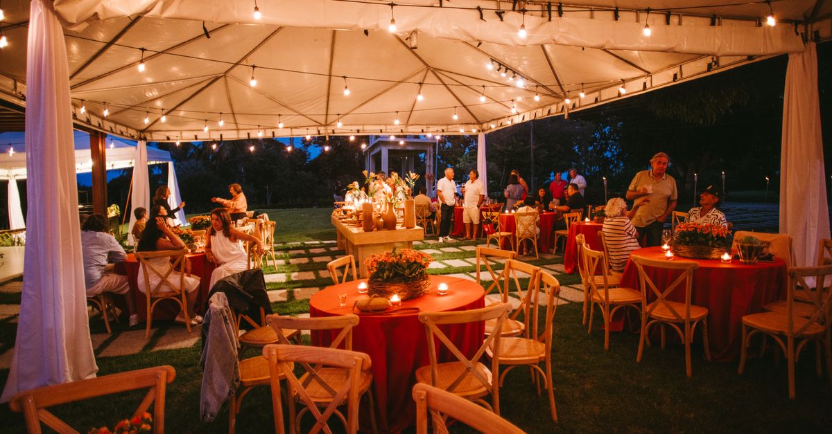 Charming outdoor evening event setup with string lights, red tablecloths, and wooden chairs under a canopy.