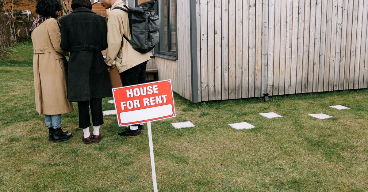 Group of adults viewing a wooden house with a 'House for Rent' sign on the lawn.