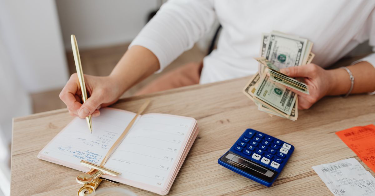 Adult holding cash and writing in planner while using a calculator at home.